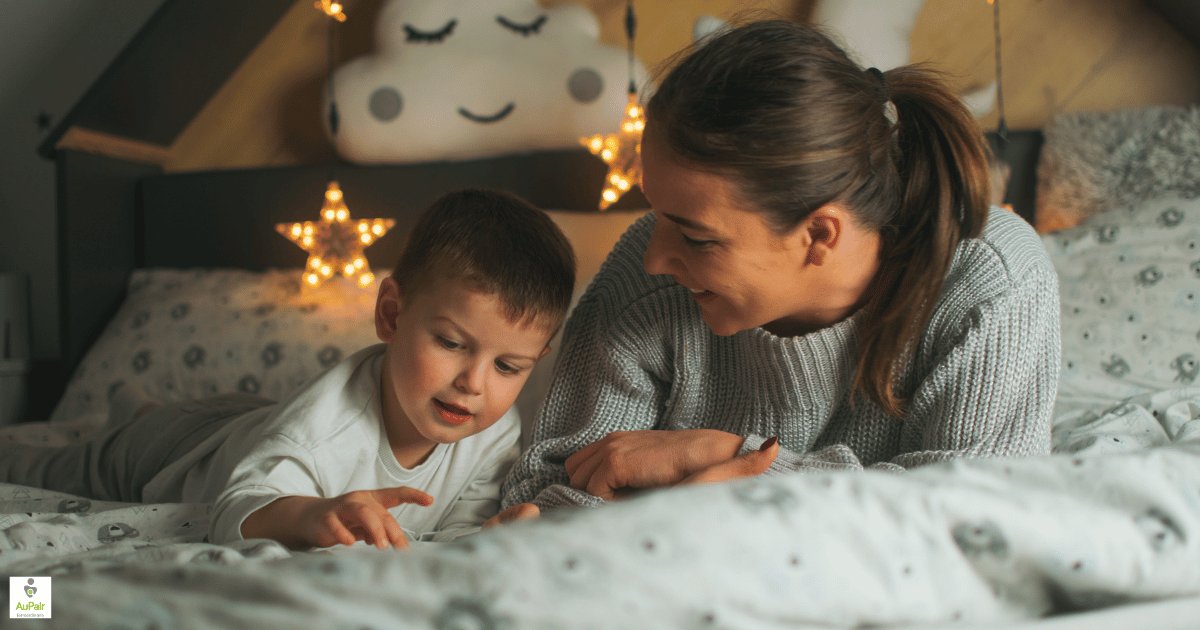 A babysitter and a young boy lying on a bed in the evening, smiling together, with star-shaped fairy lights behind them